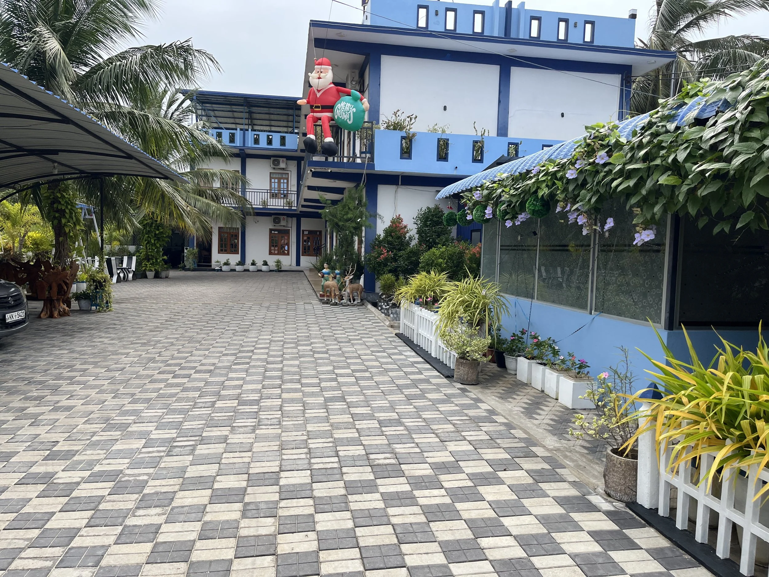 A blue and white building decorated for Christmas with an inflatable Santa Claus sitting on a balcony holding a green sack that says 'Merry Christmas', surrounded by tropical plants and trees, a car, and a paved courtyard.
