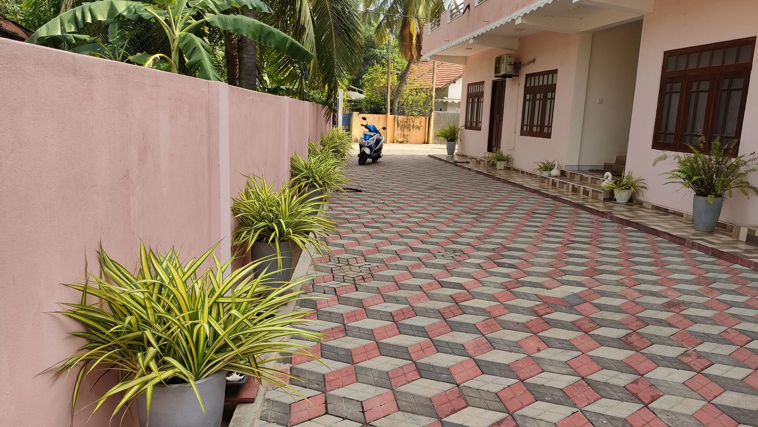 View of a sandy-colored house with brown window frames, plants in pots, a pink wall, a patterned paving driveway, and a parked scooter in the driveway.