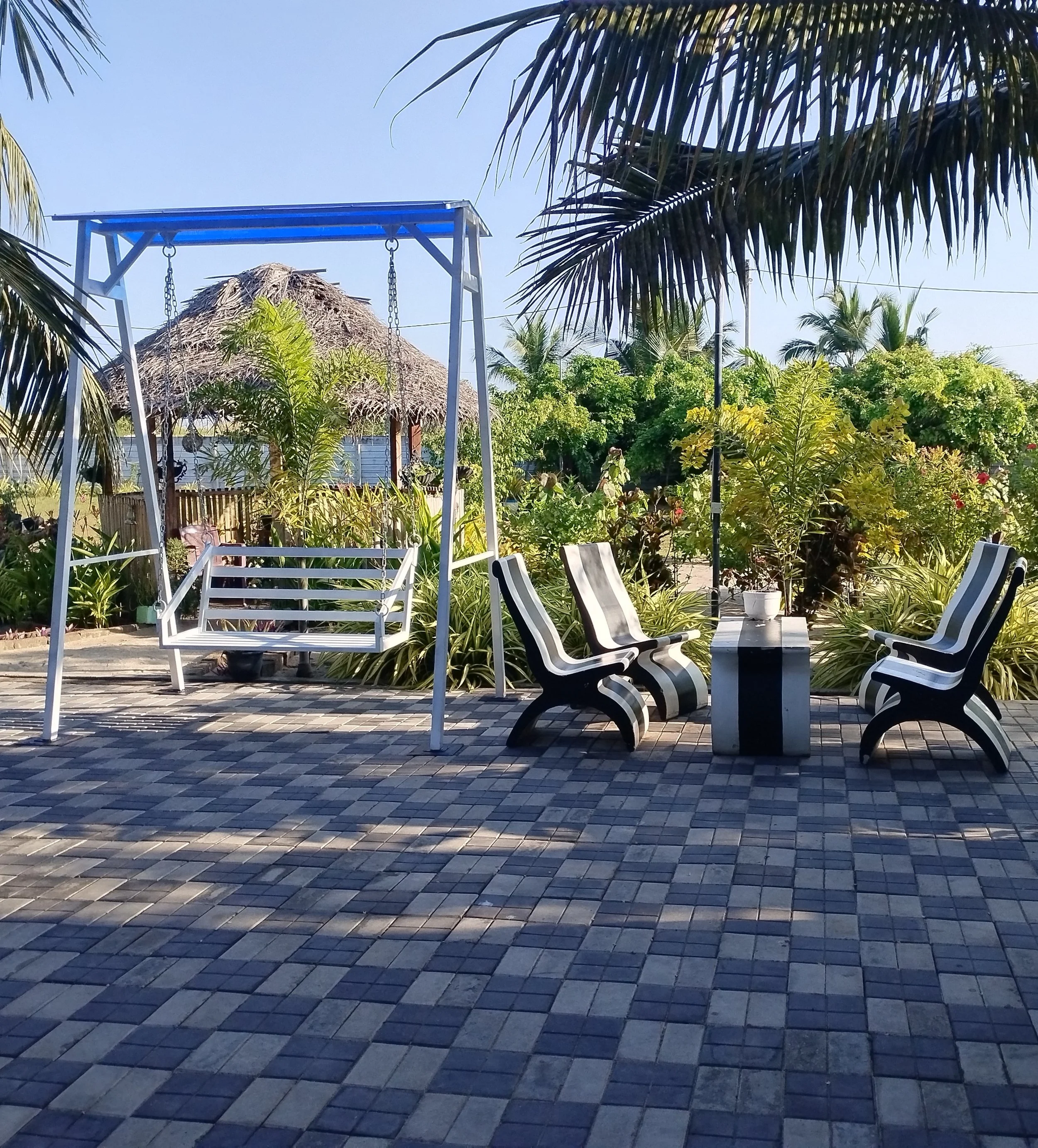 A backyard with a wooden swing set and several white chairs on a tiled patio, surrounded by lush green plants and palm trees under a clear blue sky.