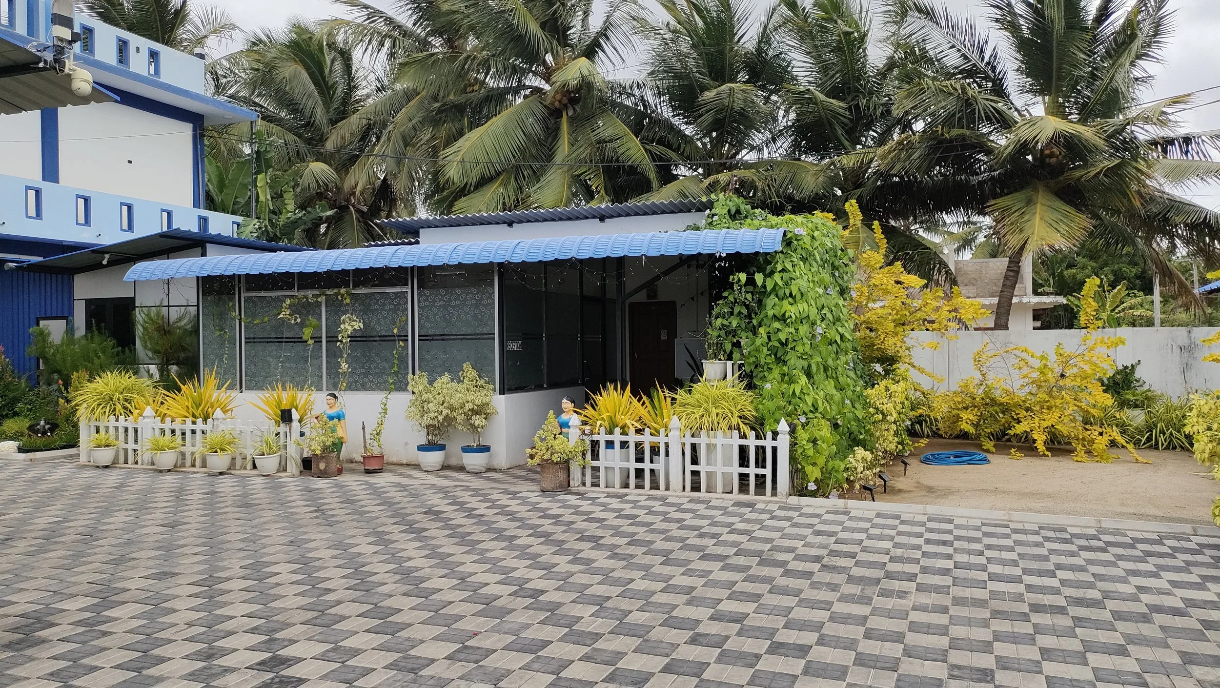View of a house surrounded by lush tropical plants and trees, with a paved front yard and a white picket fence, under a cloudy sky.