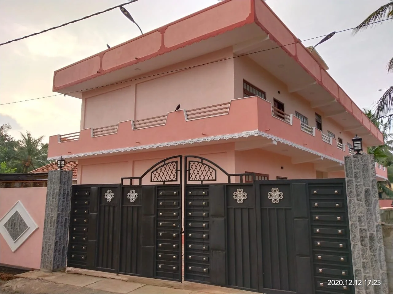 Pink multi-story house with balconies and black gate in front, surrounded by palm trees.