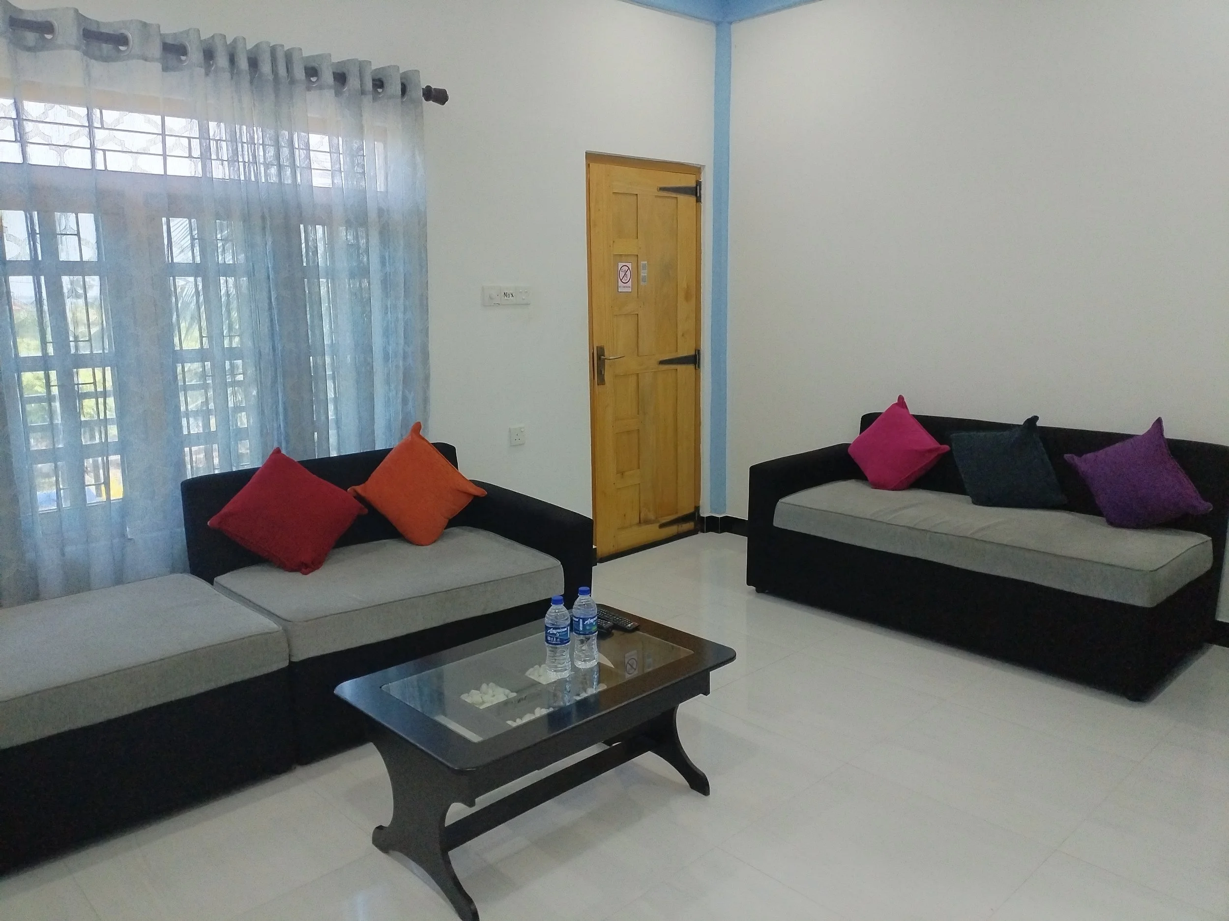 Living room with two black sofas with red, orange, purple, and black cushions, a glass coffee table with bottles of water and tissues, a window with sheer curtains, a wooden door, and a blue wall with white paint, daytime setting.