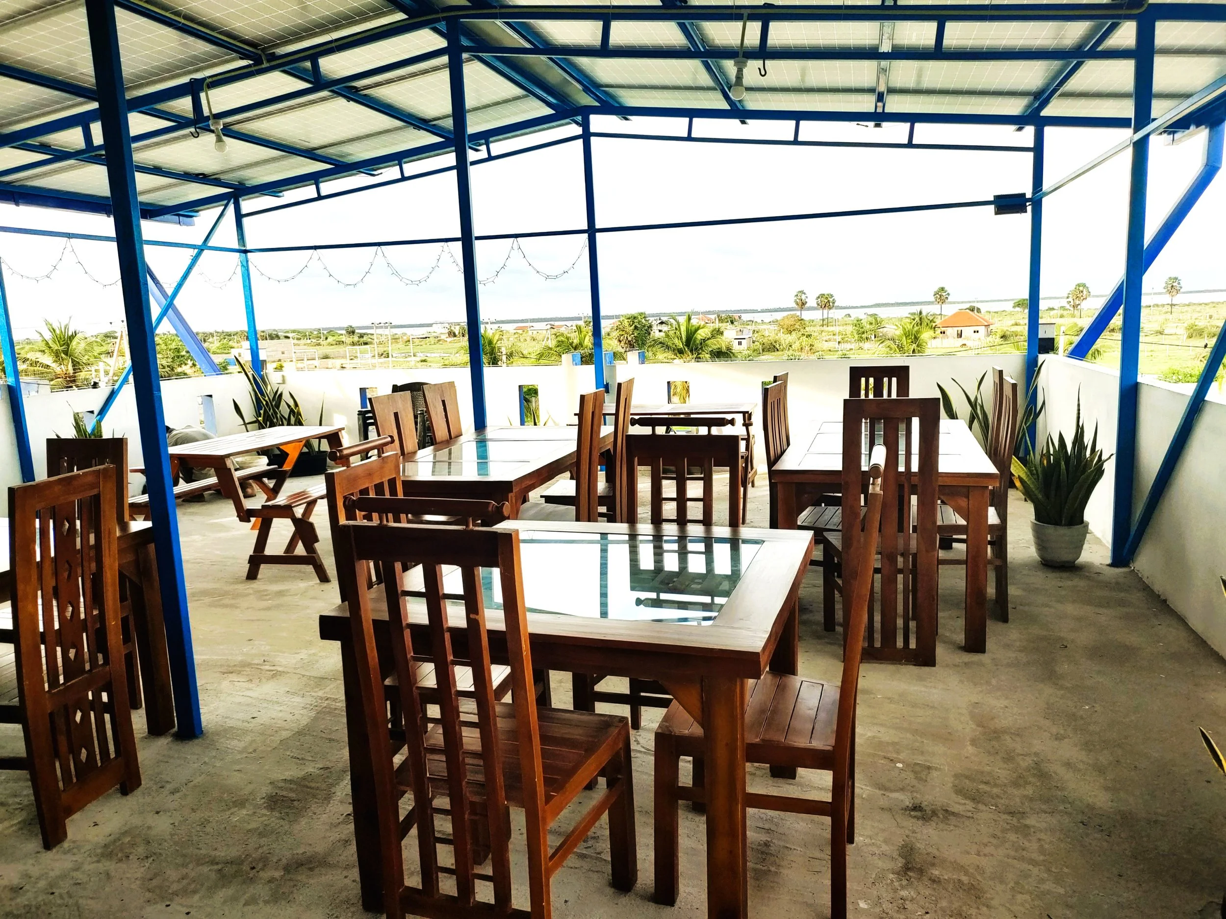 A covered outdoor terrace with wooden tables and chairs. The roof has solar panels and string lights hanging. The background shows a landscape with palm trees and a few houses under a bright sky.