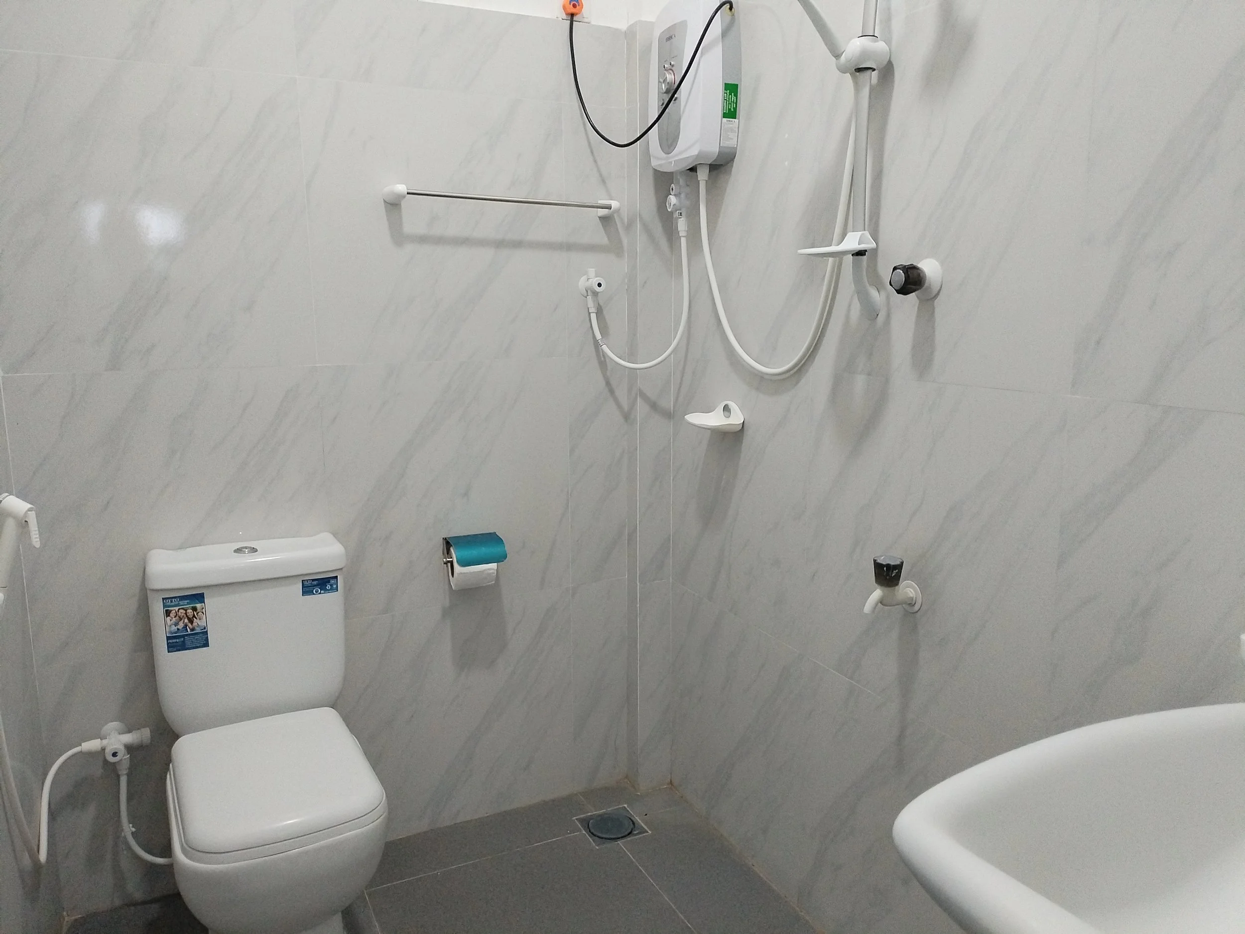 A bathroom with a toilet, a bidet, a shower area with a water heater, a grab bar, and various plumbing fixtures on light-colored tiled walls.
