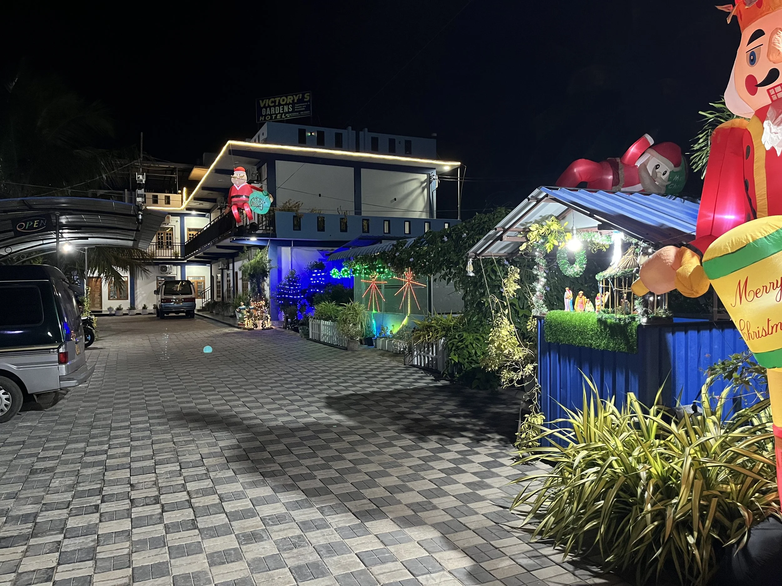 Night view of a building decorated with Christmas lights and inflated holiday figures, including Santa Claus and a reindeer, with parked cars in the foreground.