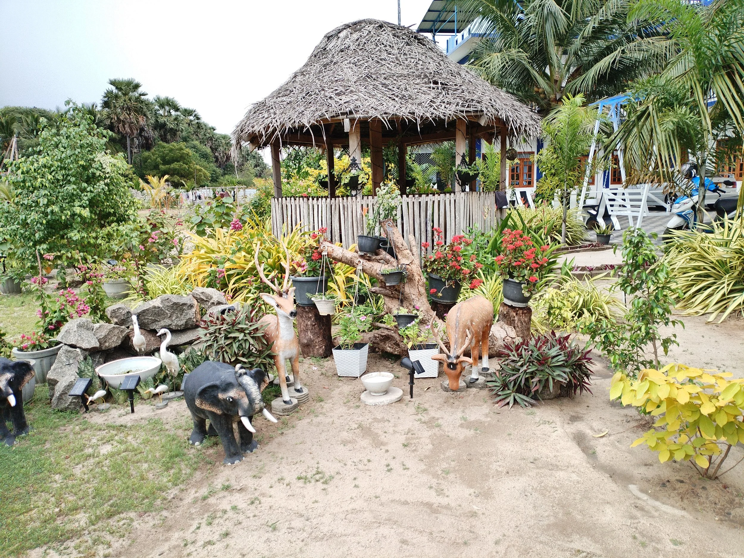 A small thatched roof hut in a lush garden area with various plants, trees, and decorative animal statues, including elephants and deer.