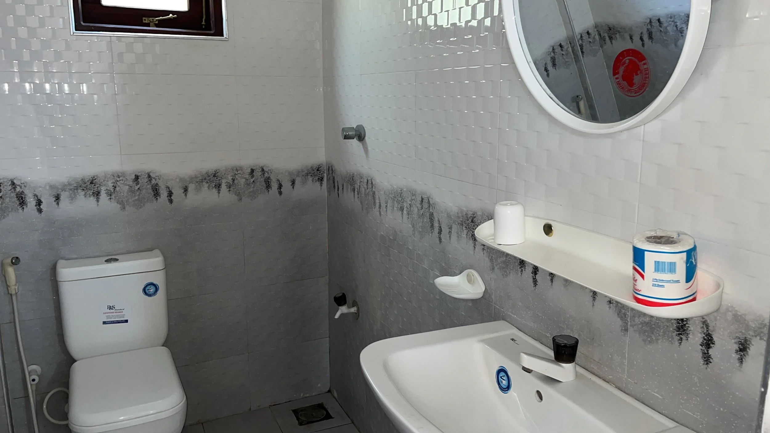 Small bathroom with white tile walls, a toilet, a bathtub, a small mirror, a toilet paper roll, and a small window. There is mold around the bottom of the walls and on the wall above the bathtub.