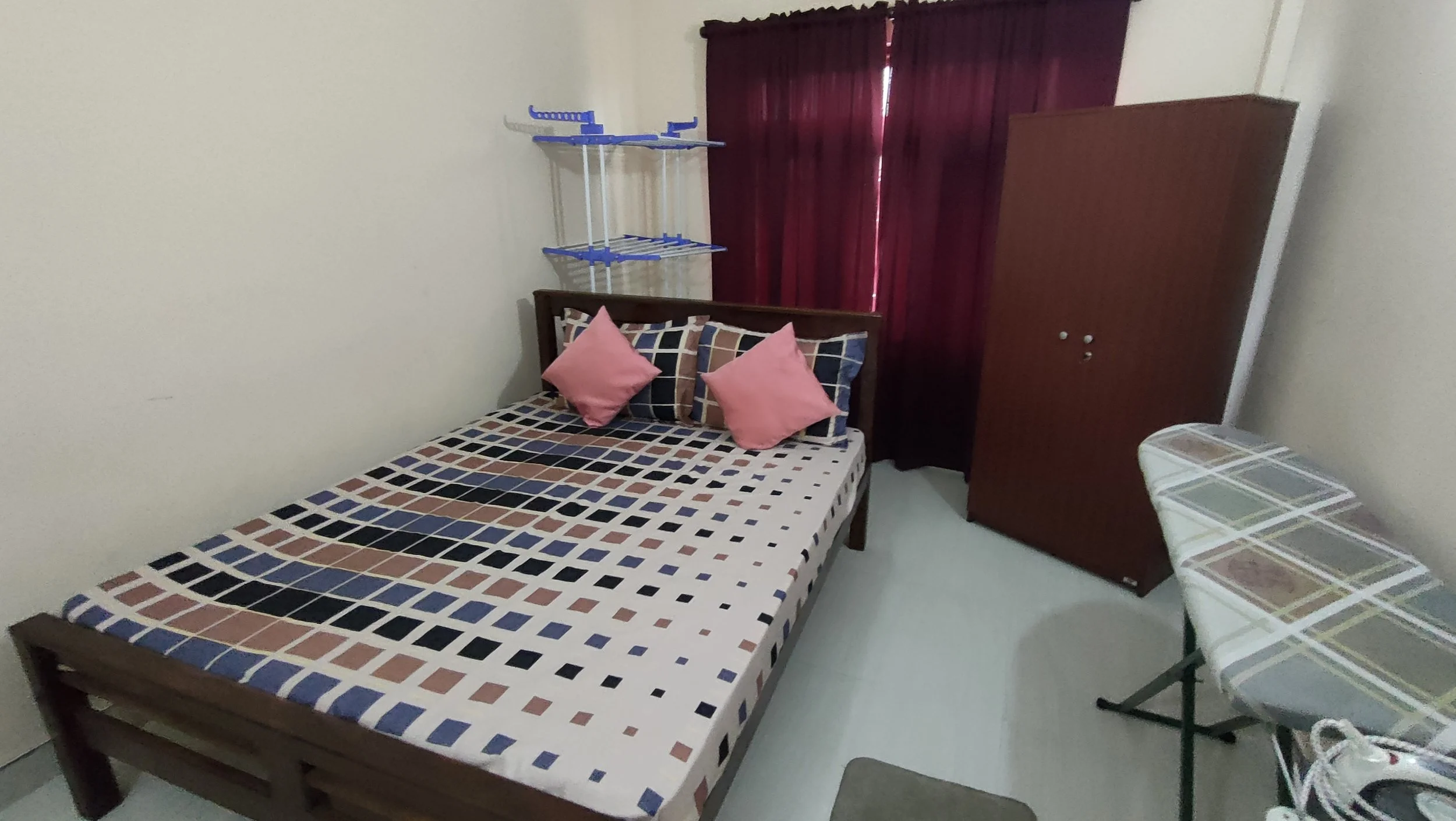 A small bedroom with a wooden bed frame and a mattress covered with a colorful checkered bedspread. Two pink pillows rest on the bed. A wardrobe stands against the wall next to red curtains on a window. There is a white plastic table with a checkered