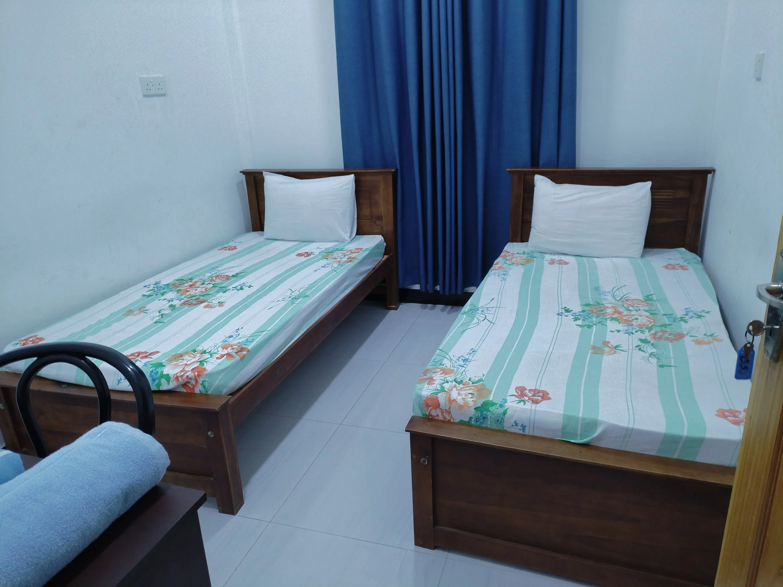 A room with two single beds, each with a pillow and floral patterned bedsheet. There are blue curtains in the background and a yellow door partially visible on the right.