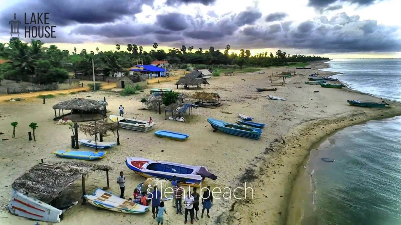 A quiet beach with boats on the sand, some huts, and a small group of people near the shoreline under a cloudy sky with dark storm clouds. The lake house logo is in the top left corner.