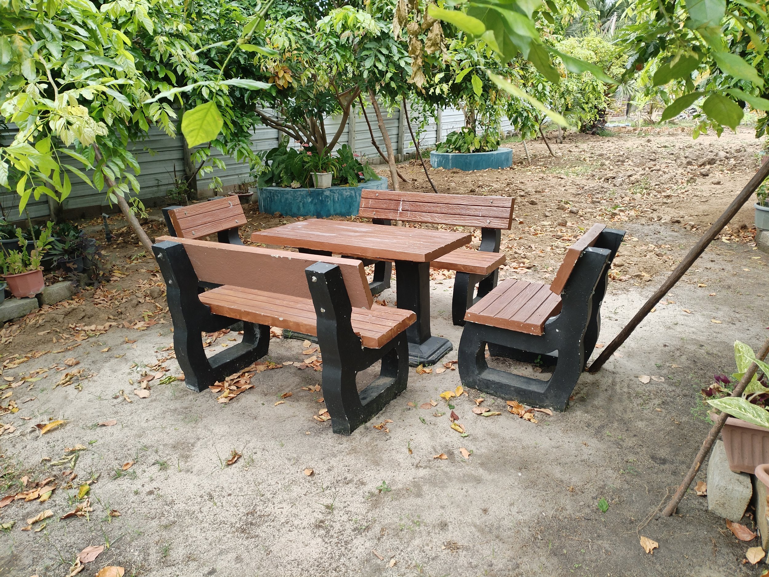 An outdoor garden scene with a wooden picnic table and benches, surrounded by green leafy trees, potted plants, and a metal fence. The ground is covered with dirt and scattered fallen leaves.