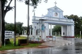 Entrance gate to a religious site with a white archway and a cross on top, surrounded by trees and a sign on the left side.
