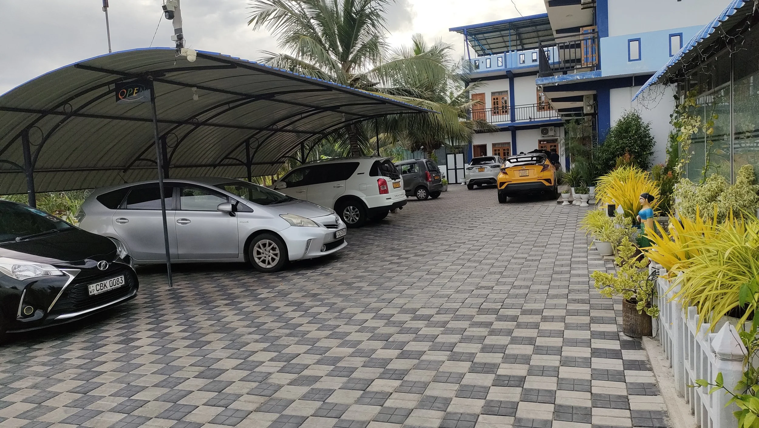 A parking lot with several cars parked under a metal shelter, surrounded by potted plants and a white fence with yellow plants on the right. A blue building with balconies and windows is in the background, with trees and a gray sky overhead.