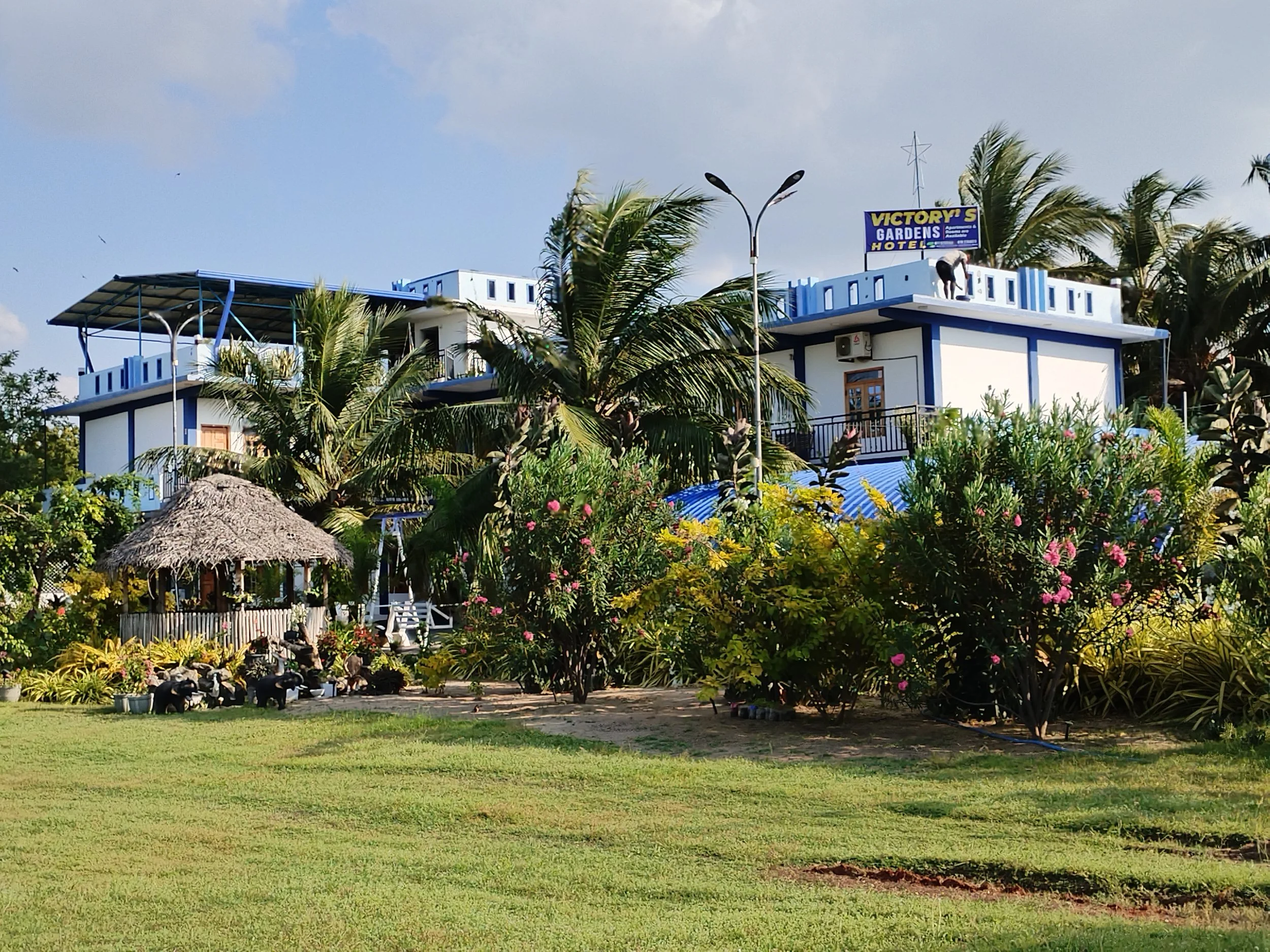 A white and blue building with multiple levels surrounded by lush vegetation, including palm trees and flowering shrubs, with a cloudy sky overhead.
