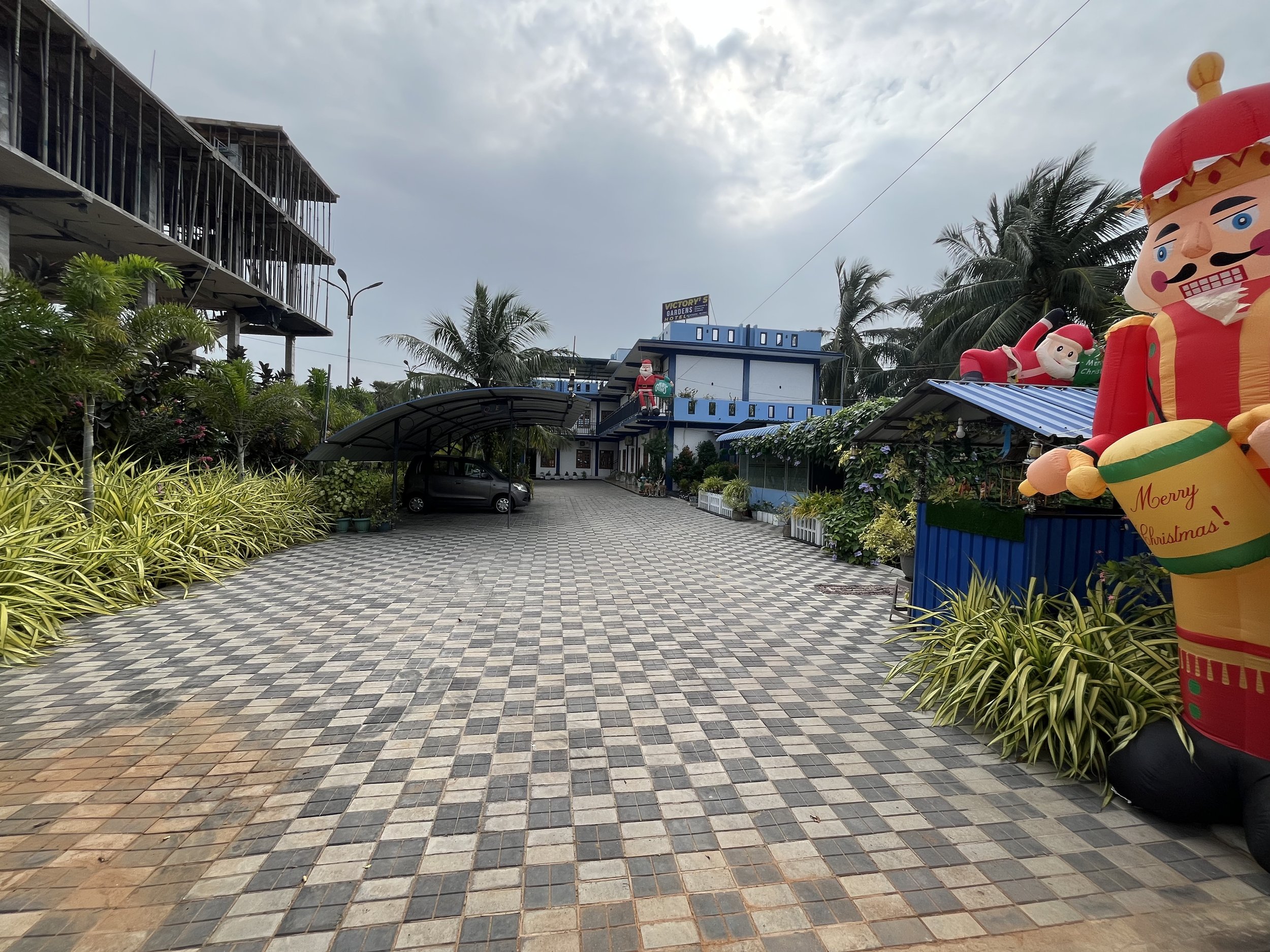Decorative Christmas inflatables including a large nutcracker figure and a Santa Claus with a green sack, set up outside in a courtyard with plants and a tiled pathway, under a cloudy sky.