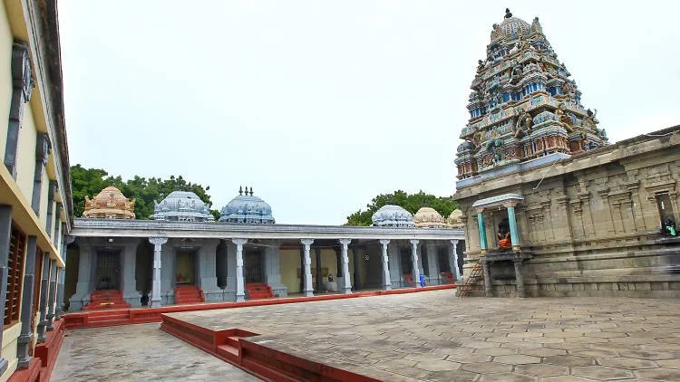 A Hindu temple with a tall, intricately decorated tower on the right and a series of white domed structures along a colonnade on the left, set in a spacious open courtyard.