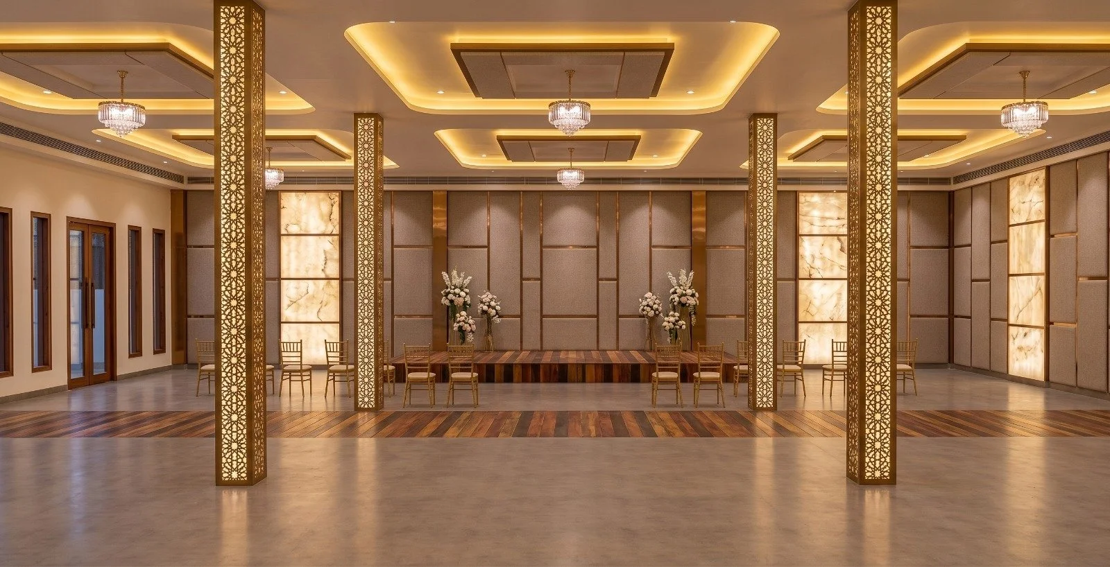 Elegant event or wedding hall with chandeliers, gold accents, floral arrangements on a low stage, and wooden and marble flooring.