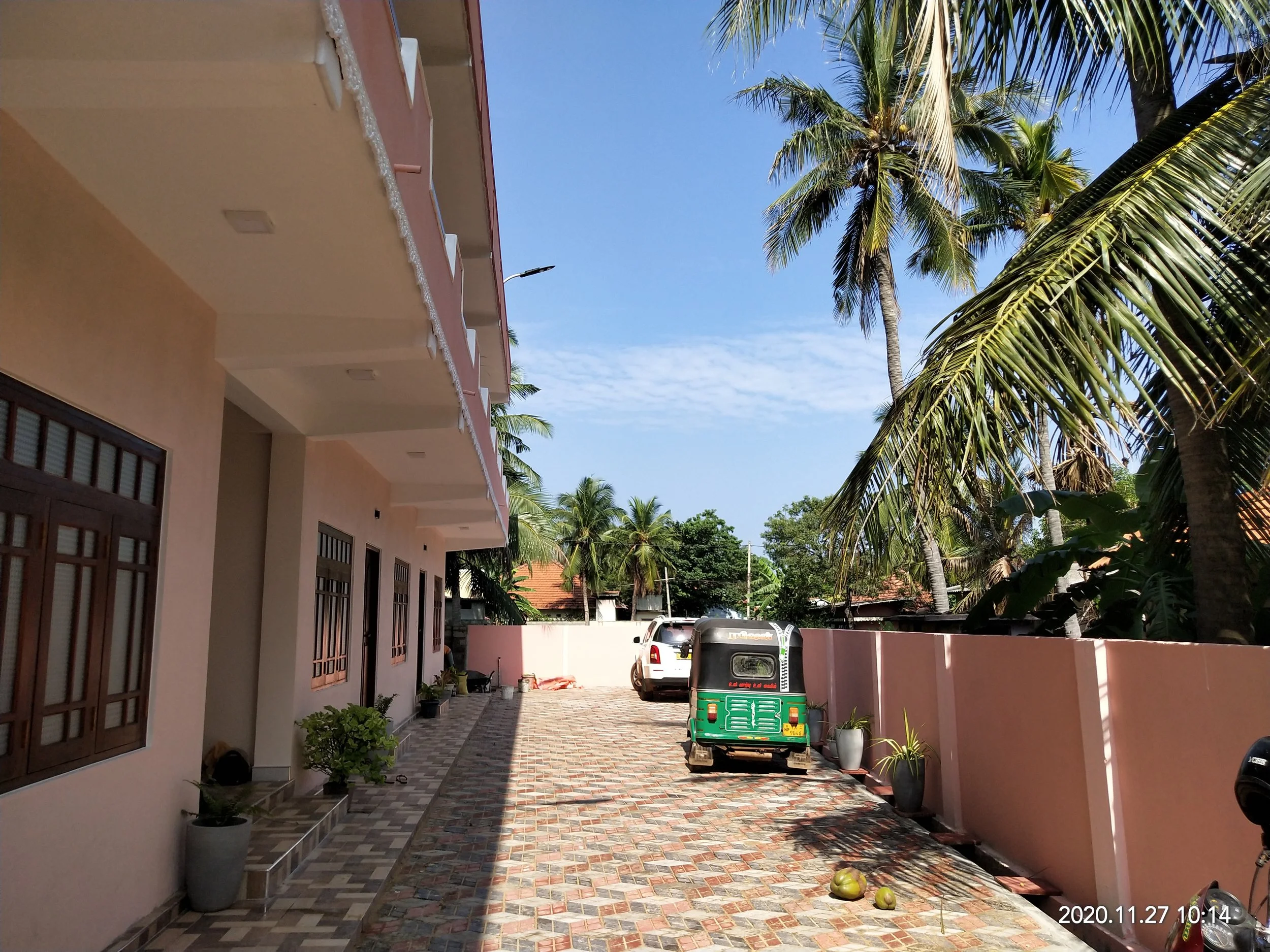 A courtyard with a peach-colored building on the left, tall palm trees on the right, and a vehicle parked in the background. Potted plants and two coconuts are on the ground. The sky is clear and blue.