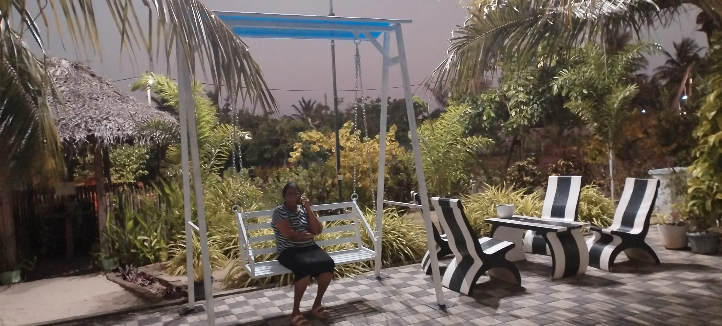 A woman sitting on a white porch swing in an outdoor garden setting during dusk, surrounded by lush green plants, palm trees, and striped black-and-white chairs with a table and bowl nearby.
