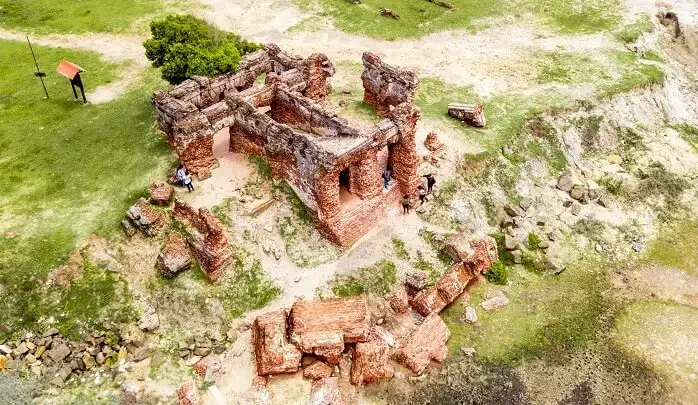 Aerial view of ancient ruins made of red bricks on grassy terrain with some stone debris and a few people exploring.