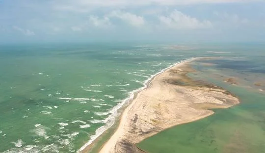 Aerial view of a coastal area with a beach, ocean waves, and a river or lagoon running parallel to the shoreline.