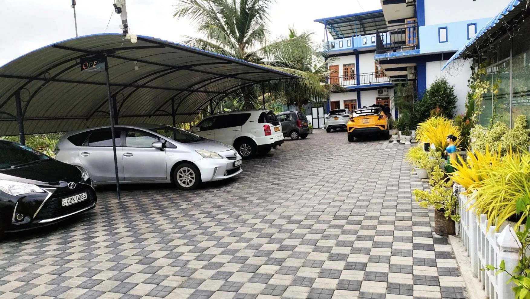 A parking lot with several cars parked, including sedans, SUVs, and a sports car, in front of a building with blue accents and lush greenery, including potted plants and decorative figures.