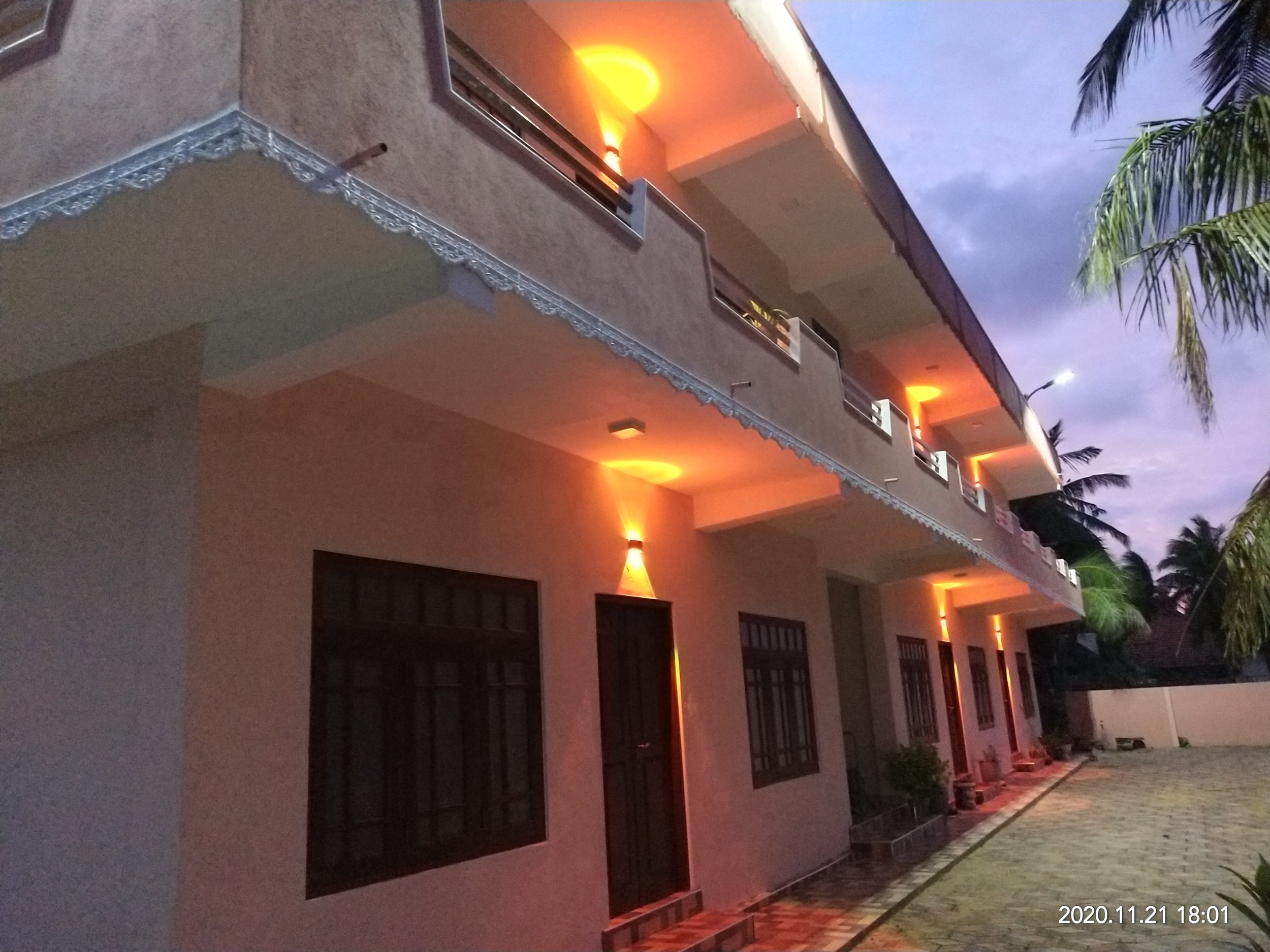 Exterior view of a two-story building with balconies, illuminated by orange and yellow wall lights, during dusk; palm trees are visible on the right side, and a paved walkway runs along the building.