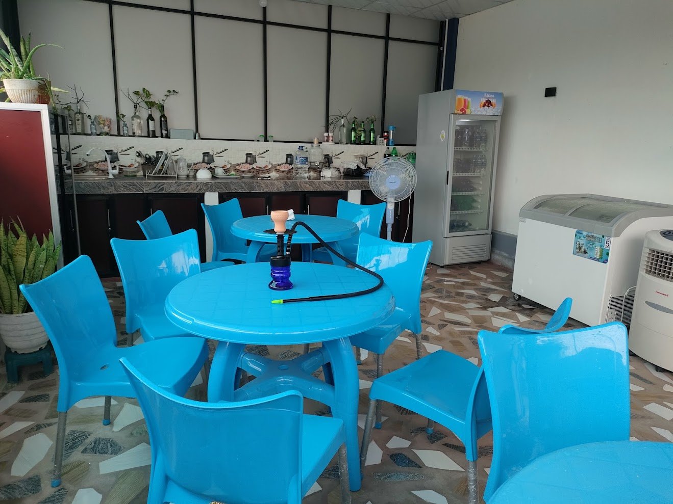 Indoor dining area with blue plastic tables and chairs, a hookah on one table, a white fan, a refrigerator, a freezer, and a water cooler, with a counter in the background