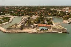 Aerial view of a historic fort surrounded by water, with a small building inside the fort, and a cityscape in the background.