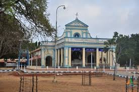 Madhu  church with a blue and white exterior, a cross at the top, and a large entrance with columns, surrounded by trees and a fenced area.