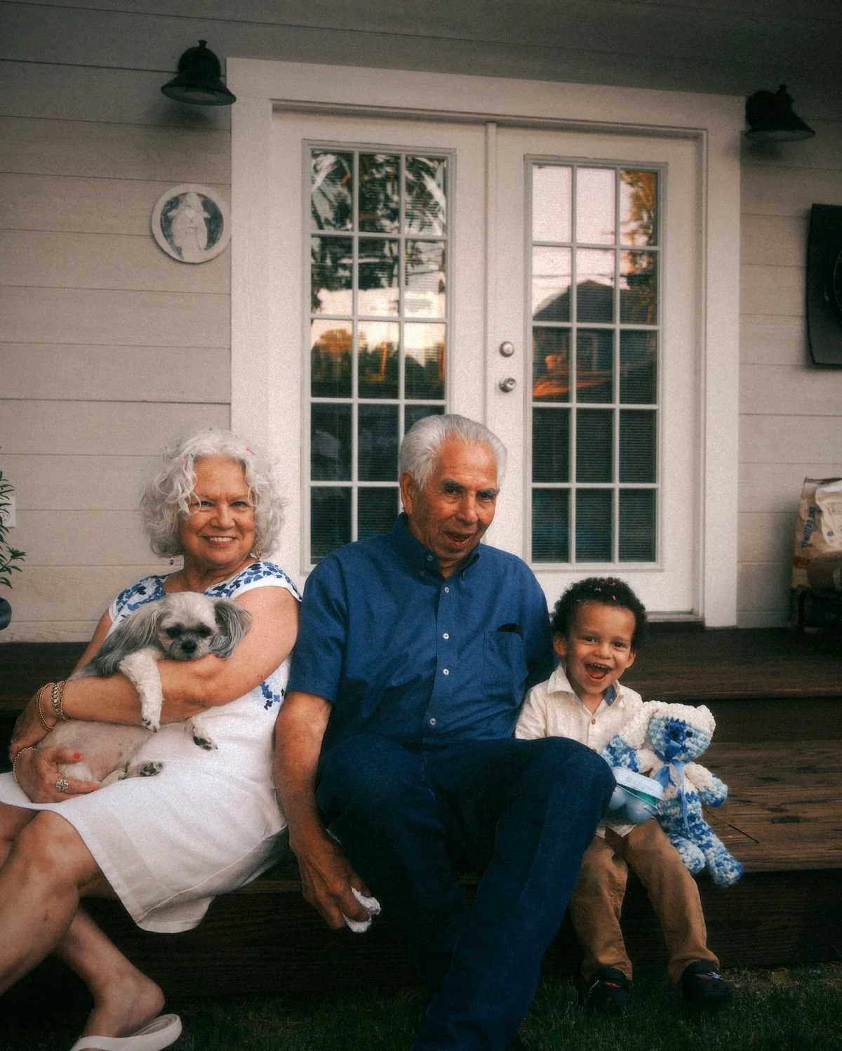 An elderly woman with white curly hair holding a small dog, sitting next to an elderly man with gray hair, and a young boy with dark hair smiling, sitting on a porch in front of a glass door.