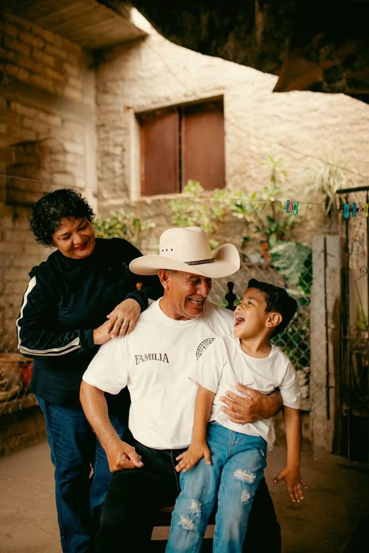 A man with a cowboy hat sitting on a chair, a woman standing beside him with her hand on his shoulder, and a young boy sitting on the man's lap. They are smiling and laughing together in a cozy outdoor setting with brick walls and greenery in the background.