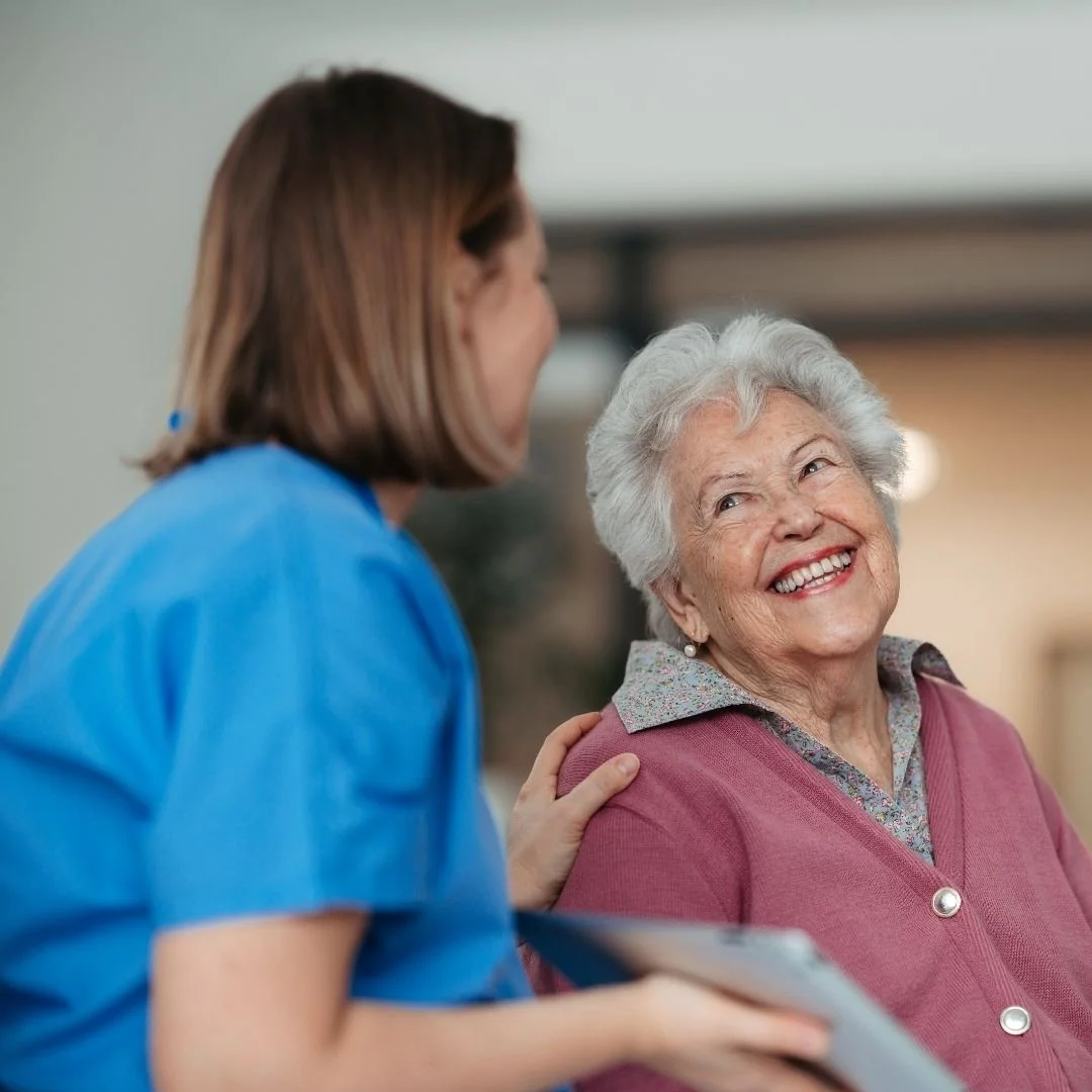 A healthcare worker in blue scrubs smiling and talking to an elderly woman with white hair, who is also smiling and wearing a pink cardigan, in a healthcare or assisted living facility.