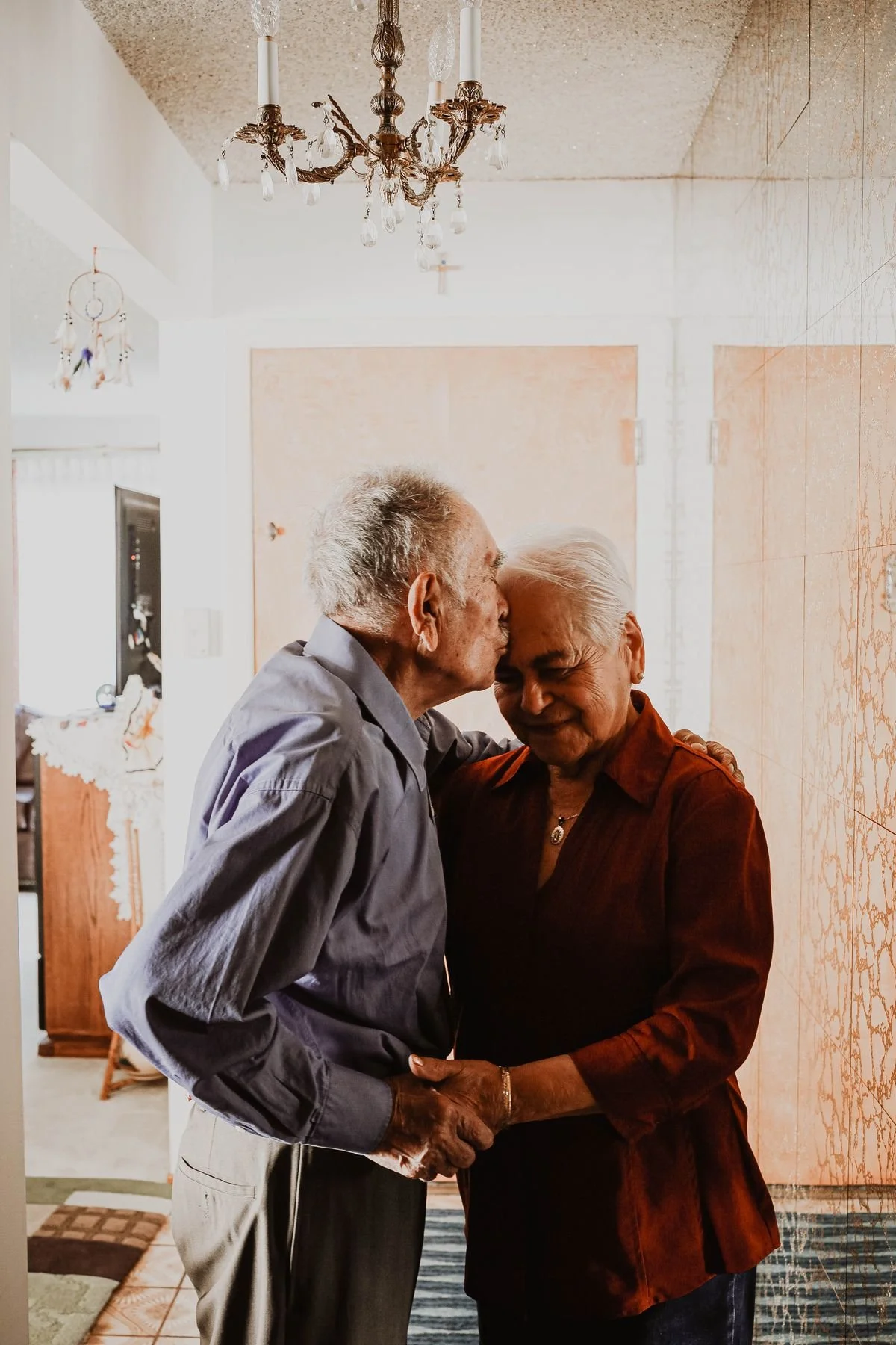 An elderly man and woman sharing a kiss on the forehead while holding hands indoors.