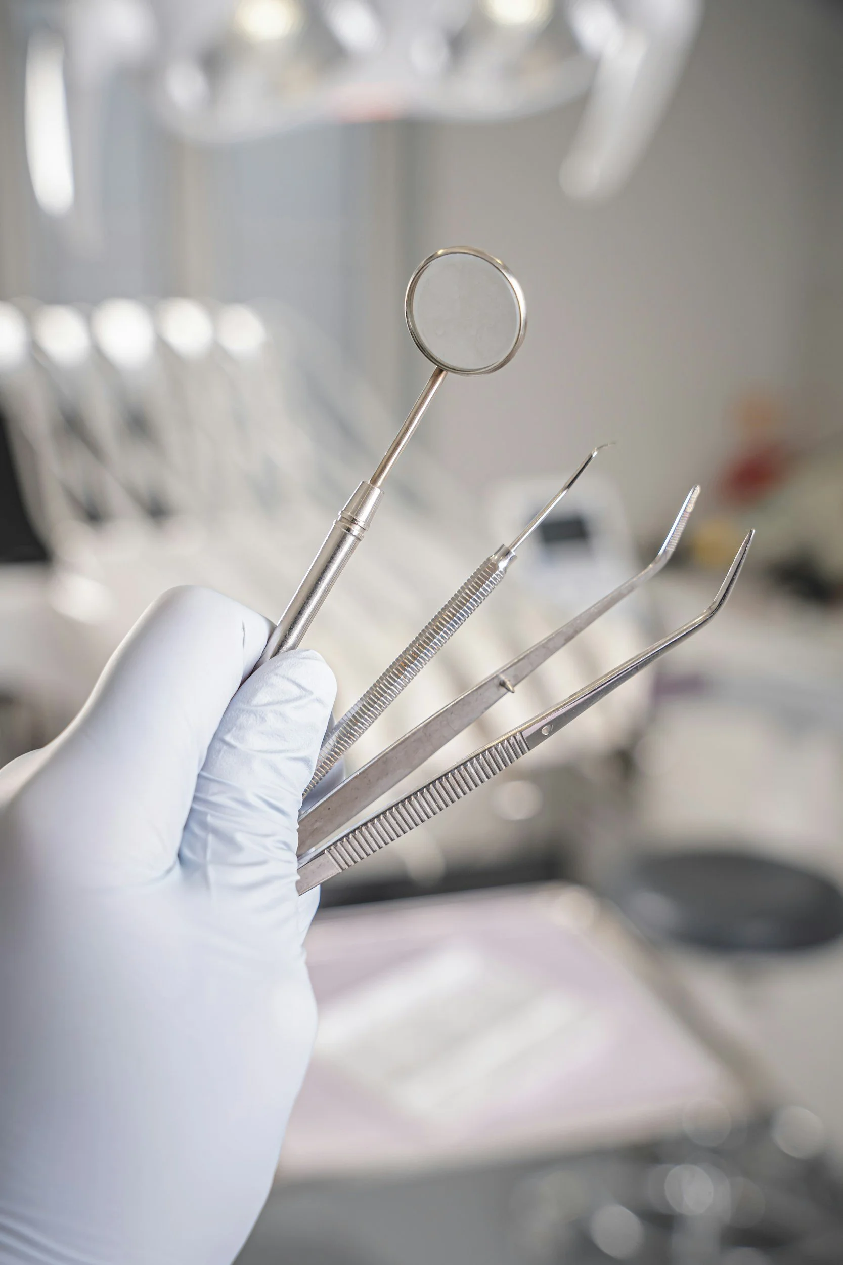 A hand wearing a white glove holds dental instruments including a mirror and other tools with metal handles, in a clinical setting.