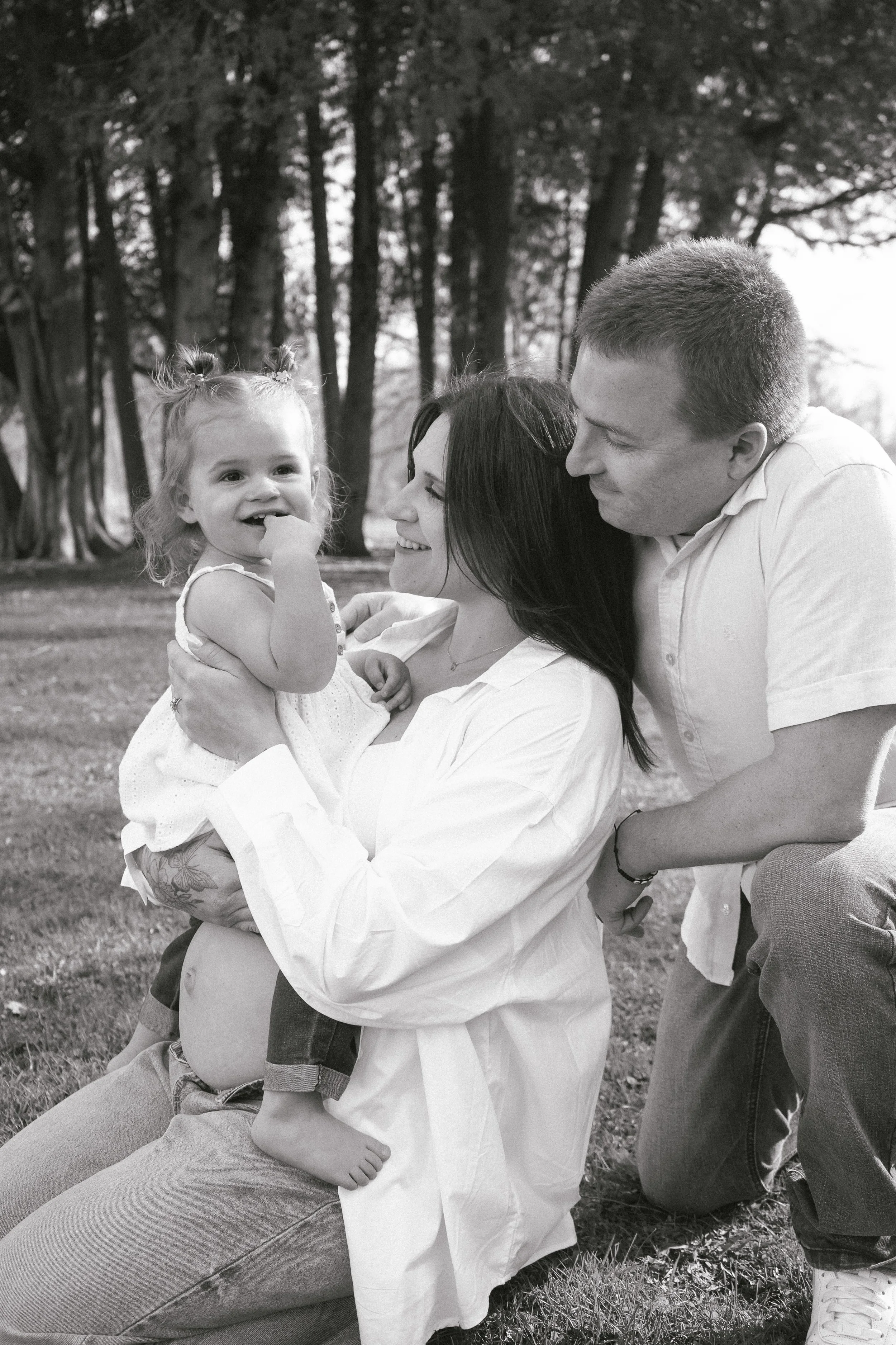 A family of four enjoying a moment outdoors in a park. The mother, father, and their young daughter are smiling and sharing a joyful interaction, with trees in the background.
