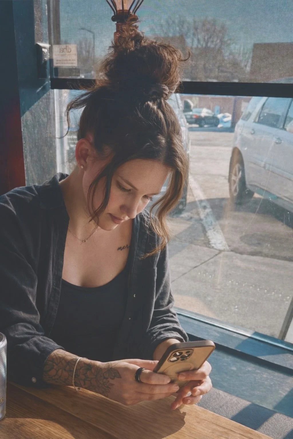 A young woman with brown hair in a messy bun, tattoos, and a black outfit, sits at a wooden table near a large window, looking at her phone in a casual indoor setting on a sunny day.
