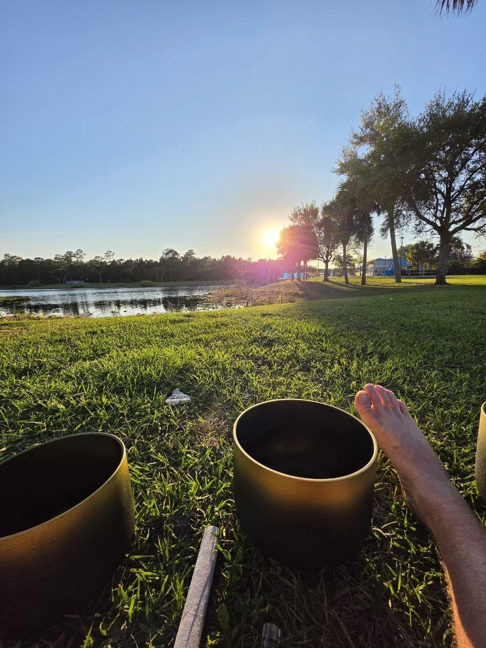 A person relaxing on grass by a lake at sunset, with music bowls and a foot resting on one of them.