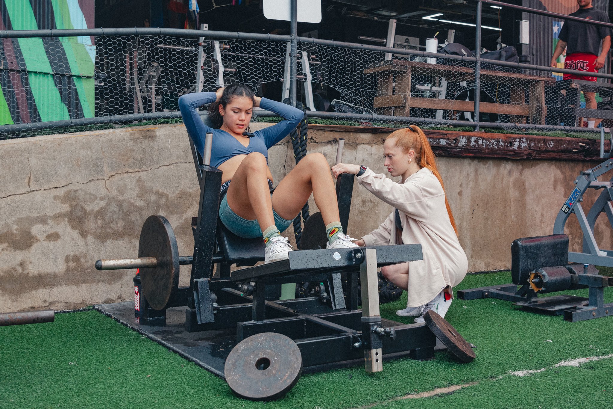 A woman with curly hair sitting on a leg press machine, doing leg exercises, while a trainer or helper with red hair and light skin assists her. The setting is an outdoor gym area with artificial grass and various workout equipment around.