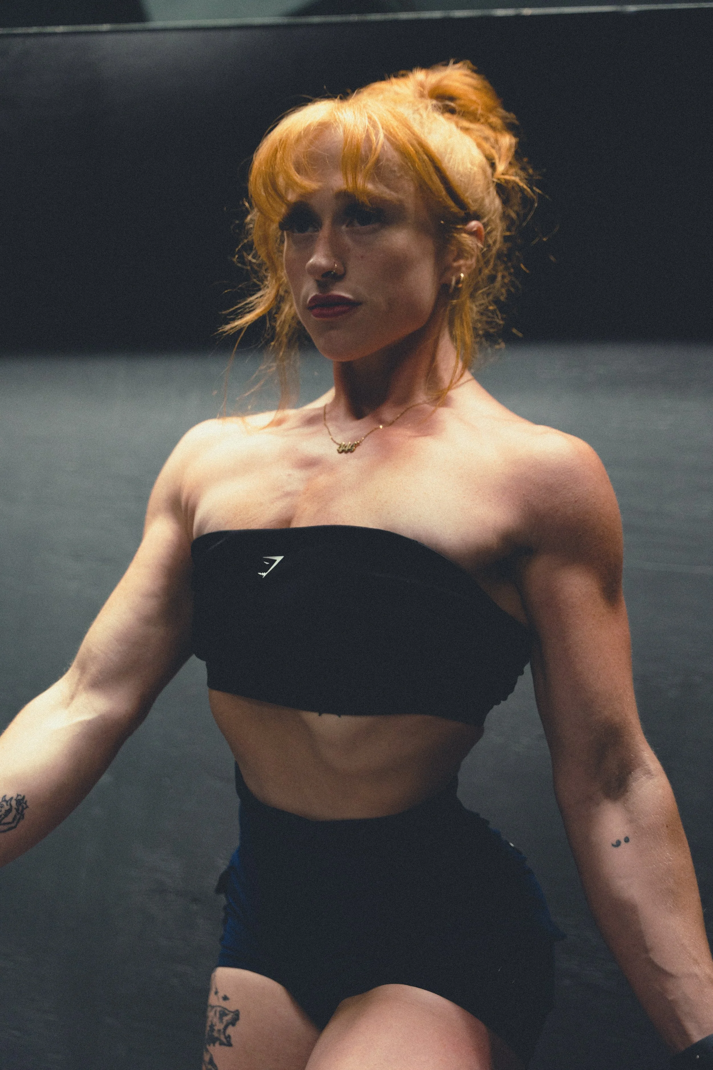 A woman with orange hair, tattoos on her arms, and a nose ring, wearing a black tube top, looking off to the side with a serious expression.