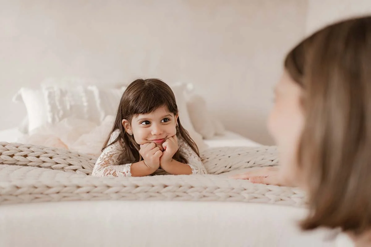 A young girl with brown hair lying on a bed, smiling, in a cozy room with a soft, light-colored background.