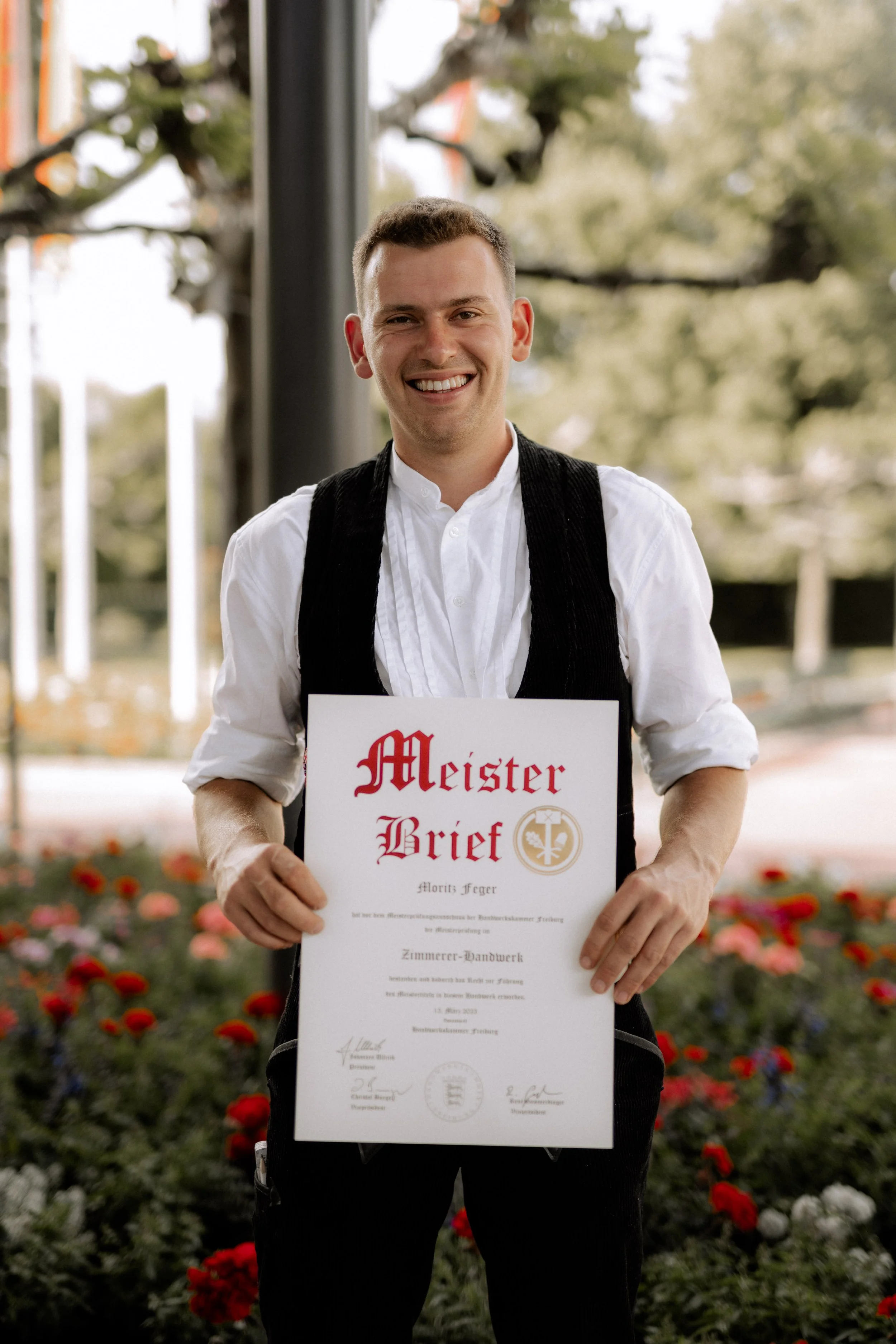 Ein junger Mann hält eine Urkunde mit der Aufschrift 'Meister Brief' und lächelt in die Kamera, im Hintergrund sind Bäume und Blumen sichtbar.