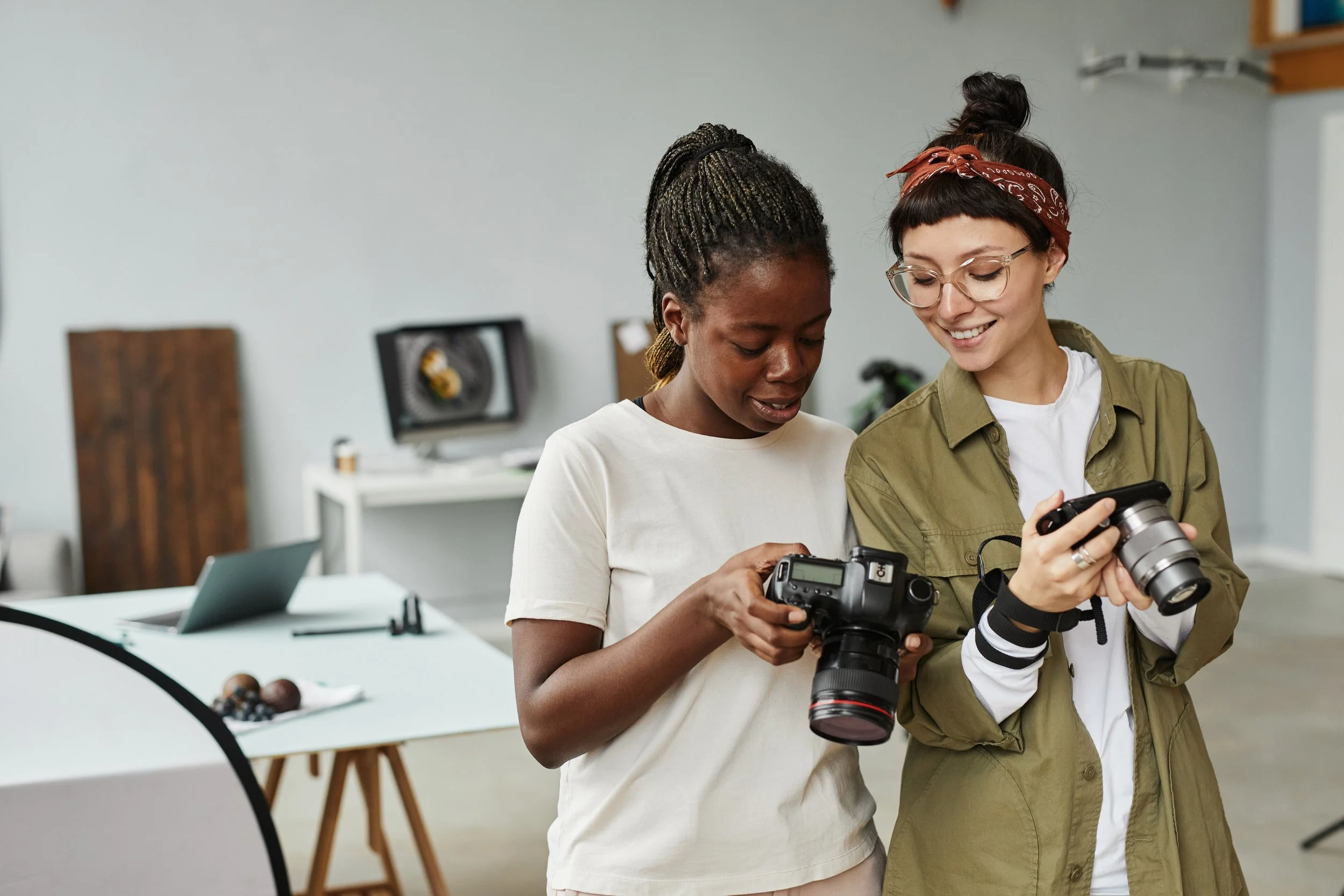 Two women standing indoors, looking at camera and phone while smiling, with photography equipment and a table with fruit in the background.