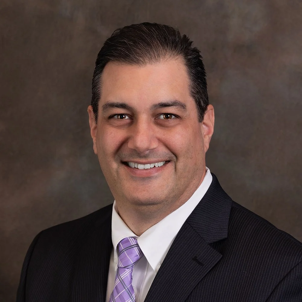 Professional headshot of a smiling man in a black suit, white shirt, and lavender striped tie.