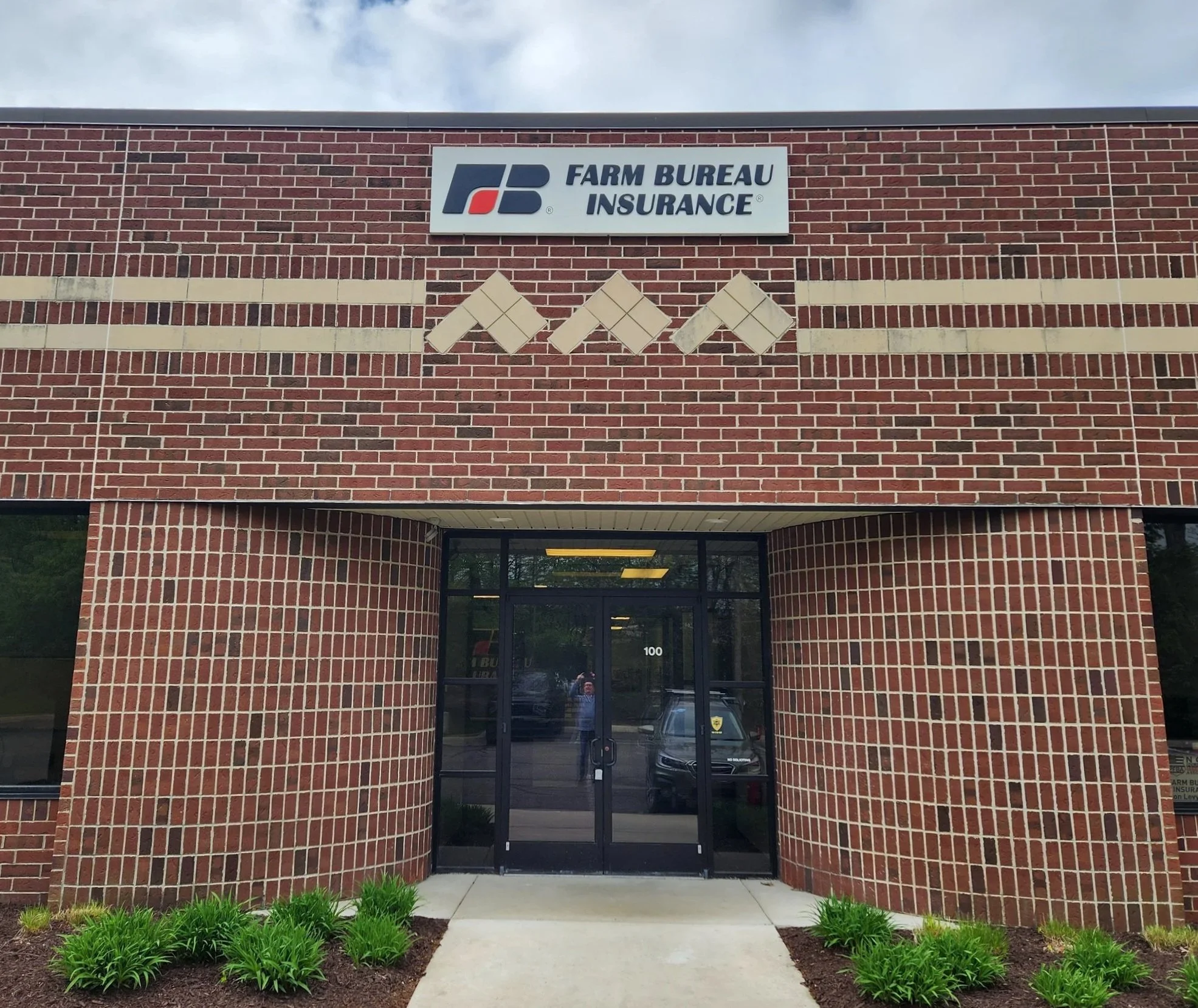 The image shows the front of a brick building with a sign that reads 'FARM BUREAU INSURANCE,' featuring the company's logo. Below the sign is an entrance door with glass panels and a decorative walkway leading to it, flanked by small green plants and grass patches.
