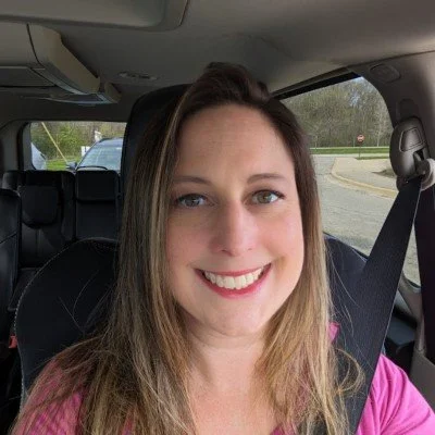 A woman smiling inside a car, with a seatbelt on, during daytime.