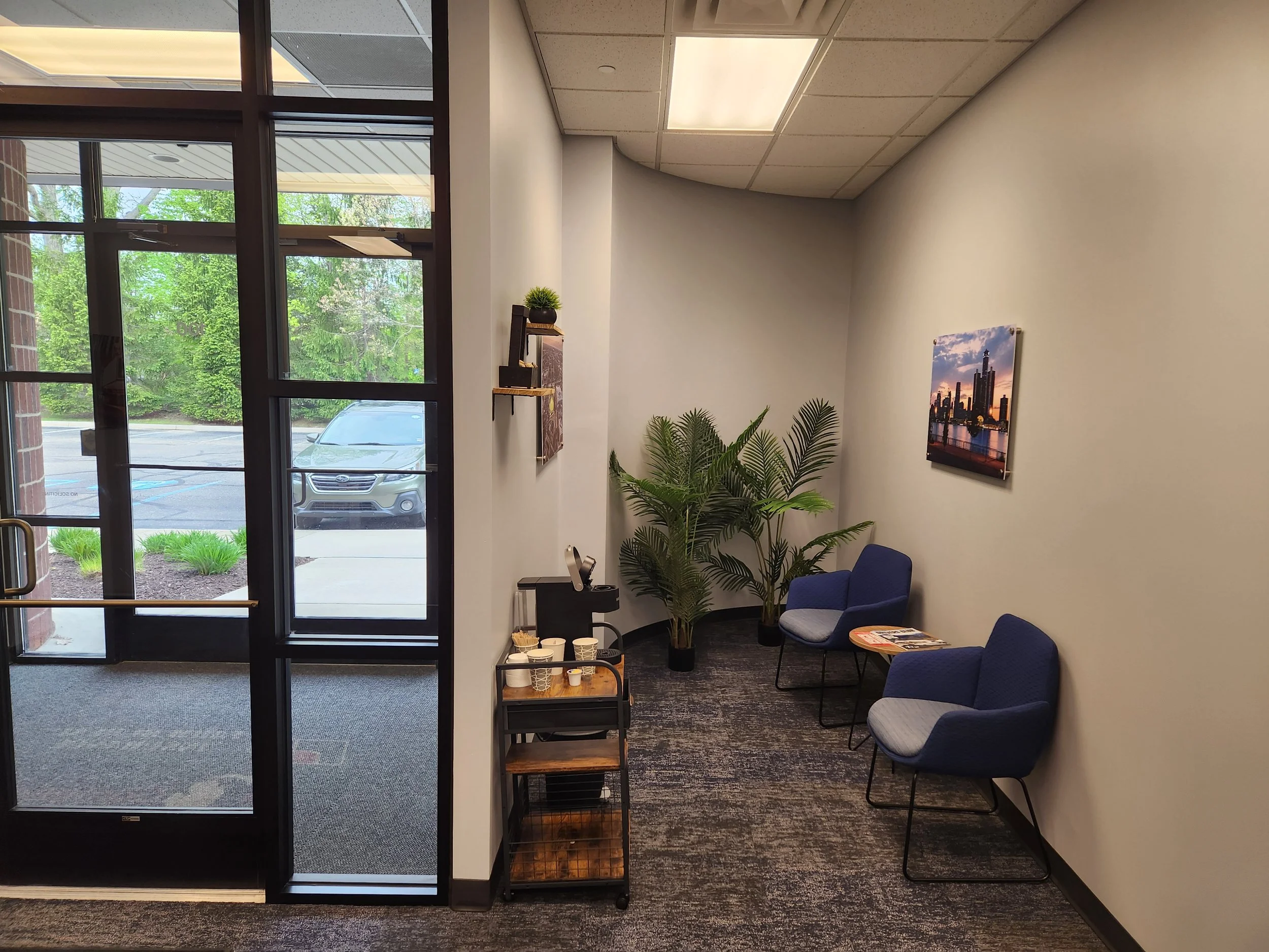 Interior of a waiting area with blue upholstered chairs, a small wooden table with magazines and disposable cups, a tall potted plant, and a wall-mounted photograph of a skyline skyline at sunset. There is a glass door and windows showing a parking lot and green trees outside.