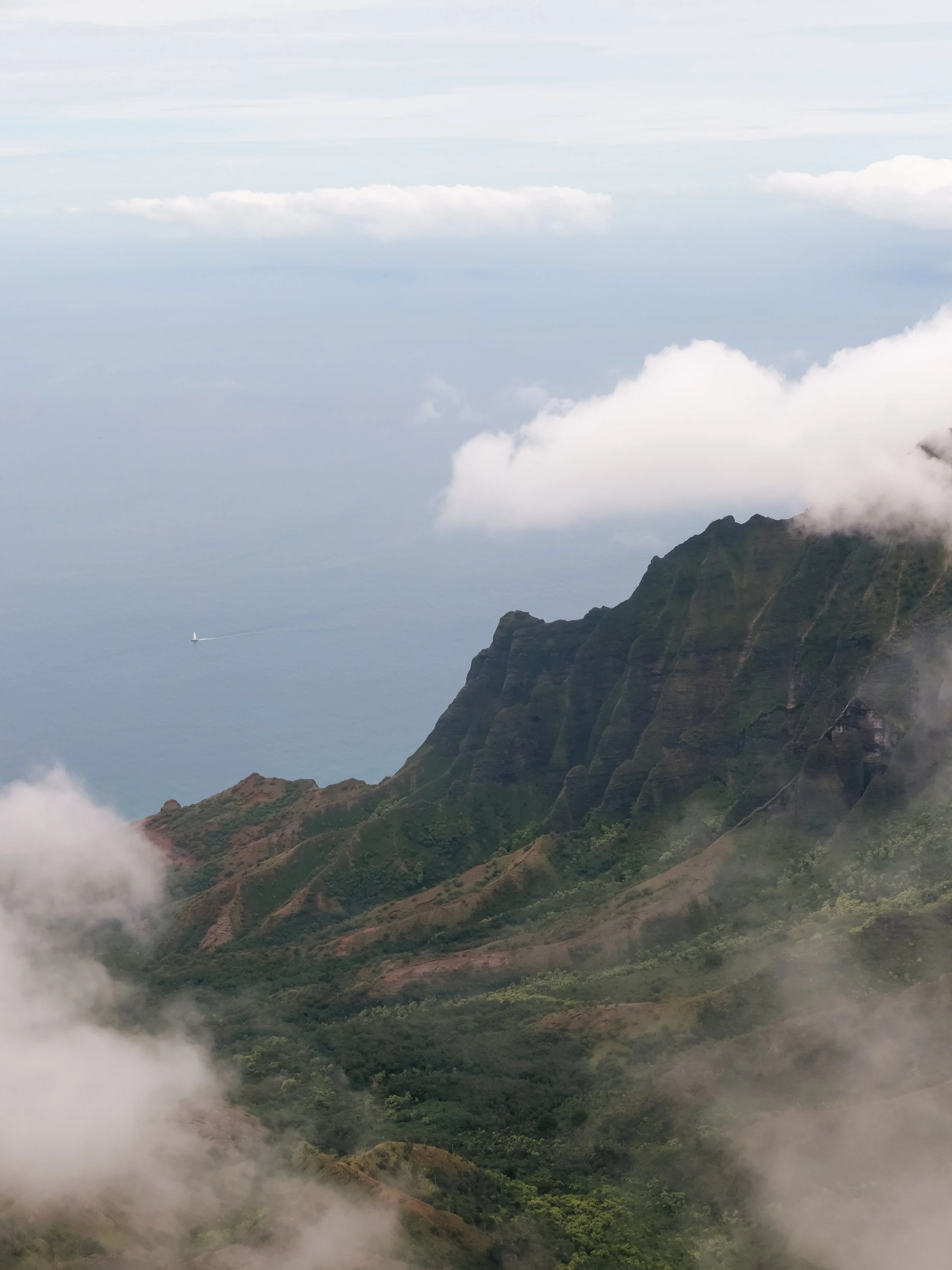 A rugged mountain range with steep, green slopes and rocky peaks, partially covered by clouds, with a sailboat visible at sea in the distance.