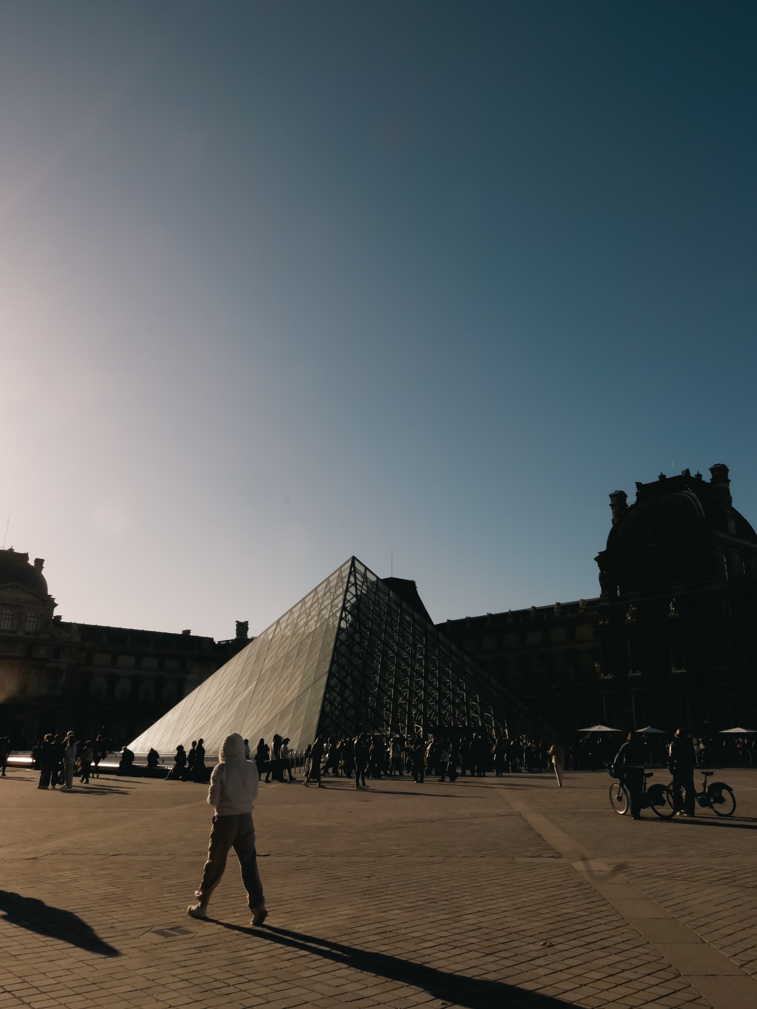 The Louvre Museum's glass pyramid entrance in Paris, with people walking and standing around outside during daylight.