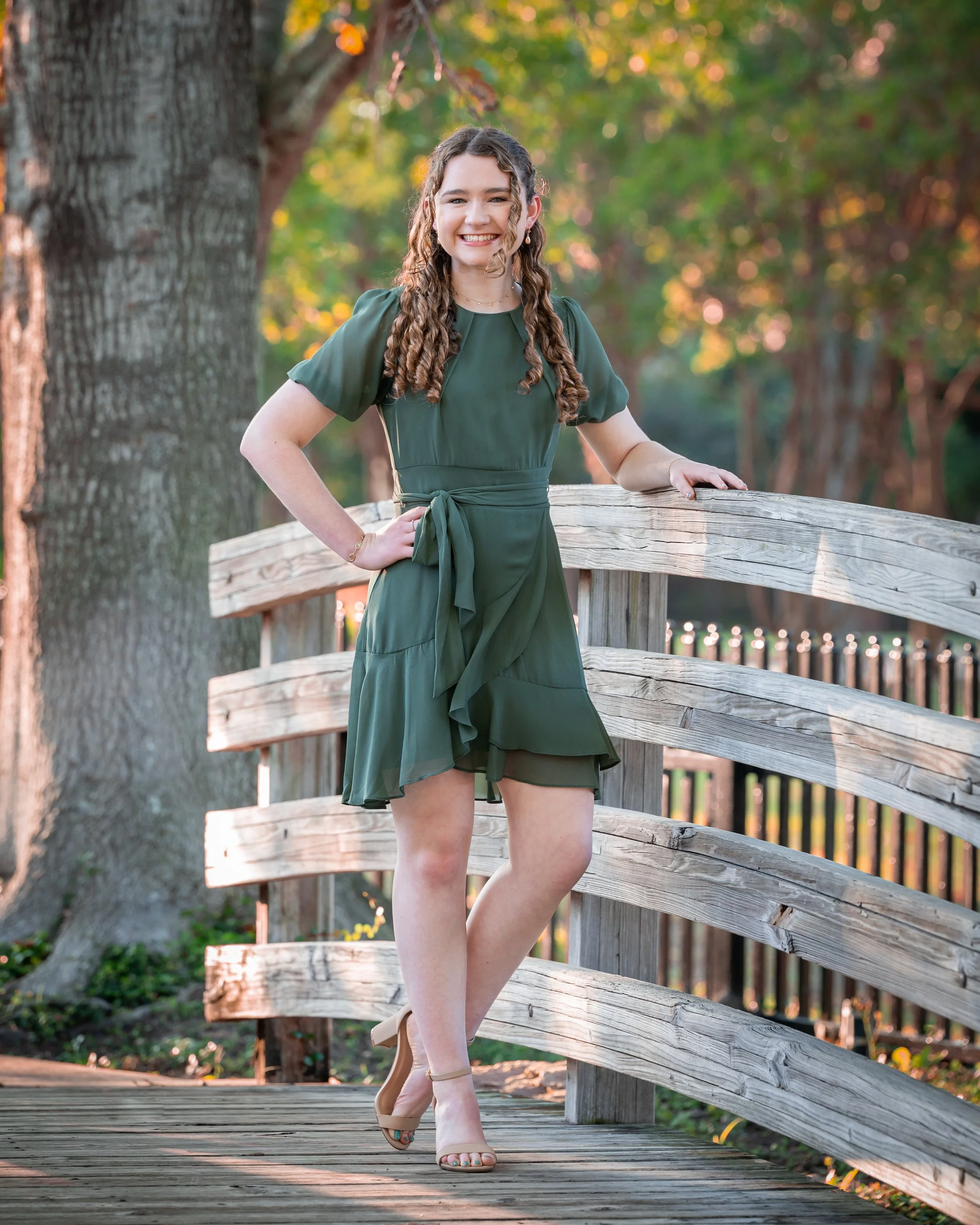 Young woman standing on a wooden bridge in a park with green trees in the background, smiling at the camera, wearing a green dress and beige high heels.